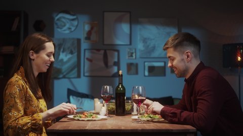 Surprised woman opening gift, a red box. Young Couple in love dinner, drinking red wine, giving present, cozy Saint Valentines Day celebration at home or in restaurant. 