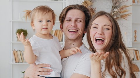 Happy caucasian family young millennial parents mother and father with little daughter infant newborn send air kiss waving goodbye smile laughing, home portrait mom, dad with baby child look at camera