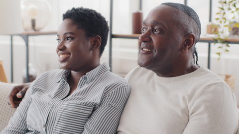 African family black married different ages, afro couple adult mature man husband and young ethnic wife sitting together on couch at home dreaming represent future ideas talking chatting about plans