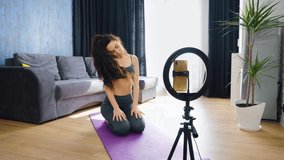 Fitness instructor sitting on knees on yoga mat and doing exercises, smartphone on tripod with light ring in front of her. Blogger recording video for online training. Concept of blogging