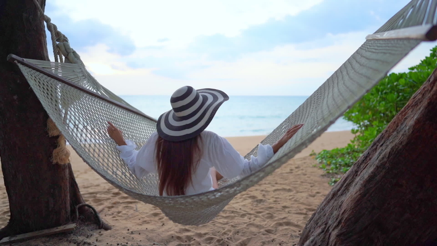 Asian woman sitting and swinging in a hammock on a tropical beach in  thailand seascape on background slow-motion back view