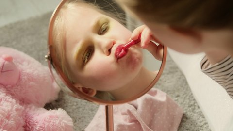 Little child girl paints her lips with lipstick, plays learn makeup in front of the mirror in the children's room. Imitates adults making experiments with cosmetics, role game.