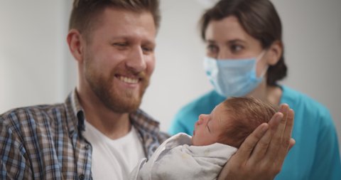 Nurse in protective mask teaching young father to hold newborn son. Happy man holding baby daughter and laughing talking to doctor in safety mask