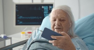 Senior female patient using earphones and smartphone for video call in hospital room. Aged sick woman lying in hospital bed and talking with family on video chat using mobile phone