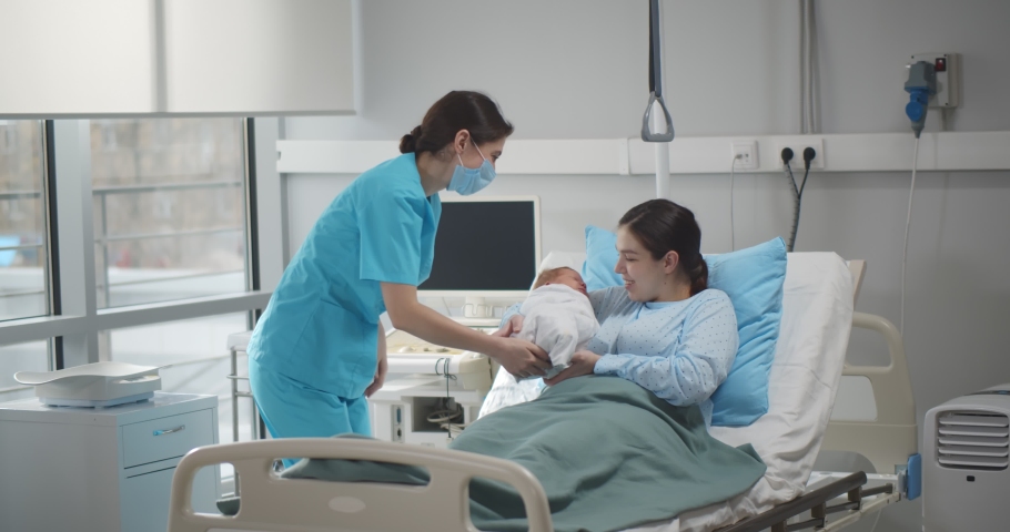 Doctor in protective mask giving newborn to young mother resting in hospital bed. Happy woman taking infant baby from nurse lying in hospital ward after childbirth Royalty-Free Stock Footage #1070798359