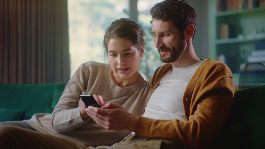 Couple Use Smartphone Device, while Sitting on a Couch in the Cozy Apartment. Boyfriend and Girlfriend Shopping on Internet, Watching Funny Videos, Use Social Media, Streaming Service. Medium Shot Royalty-Free Stock Footage #1070800651