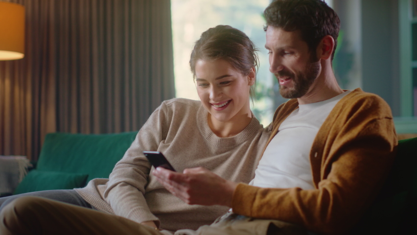 Couple Use Smartphone Device, while Sitting on a Couch in the Cozy Apartment. Boyfriend and Girlfriend Shopping on Internet, Watching Funny Videos, Use Social Media, Streaming Service. Medium Shot Royalty-Free Stock Footage #1070800651