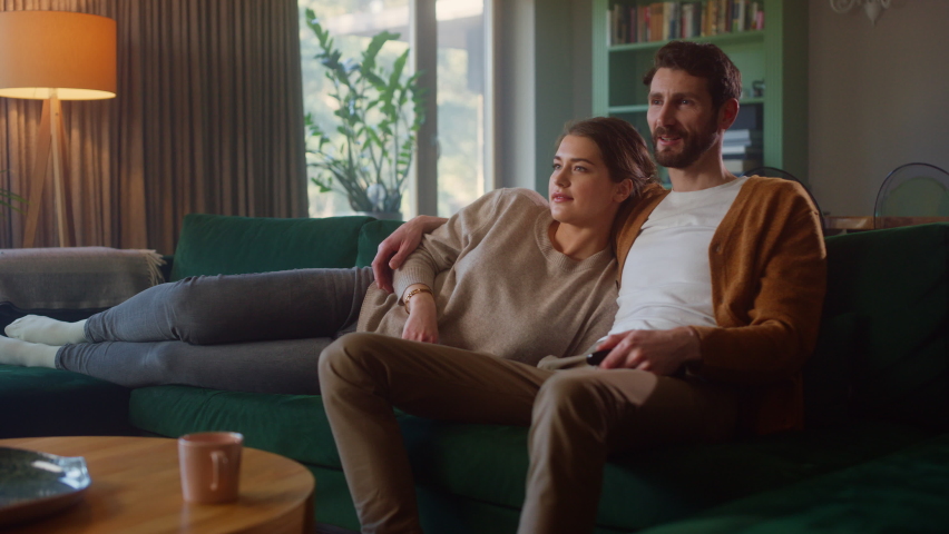 Couple Watches TV together while Sitting on a Couch in the Living Room. Girlfriend and Boyfriend embrace, cuddle, talk, smile and watch Television Streaming Services. Home with Cozy Stylish Interior. | Shutterstock HD Video #1070800663
