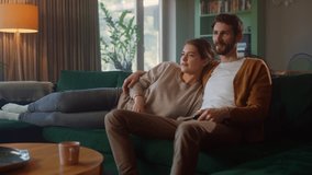 Couple Watches TV together while Sitting on a Couch in the Living Room. Girlfriend and Boyfriend embrace, cuddle, talk, smile and watch Television Streaming Services. Home with Cozy Stylish Interior.