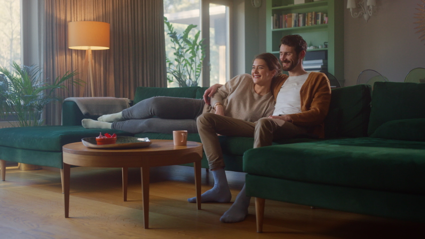 Couple Watches TV together while Sitting on a Couch in the Living Room. Girlfriend and Boyfriend embrace, cuddle, talk, smile and watch Television Streaming Services. Home with Cozy Stylish Interior. Royalty-Free Stock Footage #1070800663