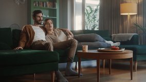 Couple Watches TV while Sitting on a Couch in the Living Room. Girlfriend and Boyfriend lying embracing, switch Channels with Remote. Spending Weekend Together at Home with Cozy Stylish Interior.