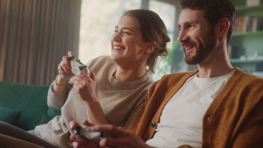 Happy Couple Sitting on the Sofa Playing Video Games, Using Controllers. Competitive Girlfriend and Boyfriend in Love have Fun Playing in Online Video Games at Home Together Royalty-Free Stock Footage #1070800768