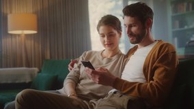 Couple Use Smartphone Device, while Sitting on a Couch in the Cozy Apartment. Boyfriend and Girlfriend Talk, do e-Shopping on Internet, Watching Funny Videos, Use Social Media, Streaming Service.
