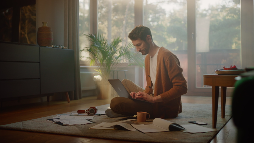 Man Using Laptop at Home, Does Remote Work. Handsome Smiling Male Sitting on the Floor Works with Papers, Documents, Brainstorms Creative Project with Research. Productive Work in Sunny Living Room Royalty-Free Stock Footage #1070800915