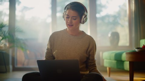 Young Woman Using Laptop at Home, Does Remote Work, Listens Music through Headphones. Beautiful Smiling Girl Sitting on the Floor Enjoys Music, Dances a Bit, Brainstorms Creative Project