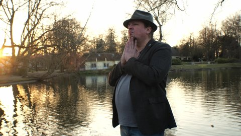 [4k] goatee man wearing hat singing an opera outdoors in front of pond in sunset