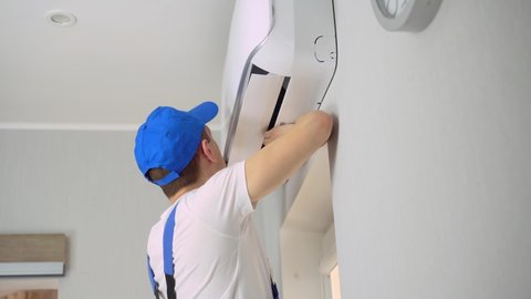 A young master in uniform, blue overalls and a cap, a white T-shirt, installs or repairs the air conditioner in the apartment room.
