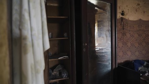 In a gloomy room of an abandoned house there is an open old wardrobe and on the shelves there is all sorts of junk. The camera moves smoothly showing the room