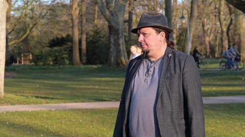 [4k] long haired middle aged man with goatee and hat is annoyed about friend comming late in public park