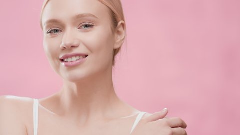 Young fit cute European woman with long golden hair in ponytail in white bikini turns her head to the camera touching her shoulder and smiling wide against pink ripple background | Skincare concept
