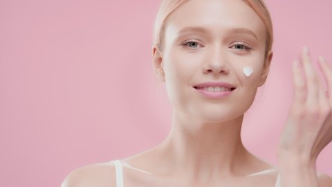 Attractive European woman with golden hair in white crop top applies moisturizer on her cheek smiling for the camera against pink ripple background | Moisturizer applying shot for face care commercial
