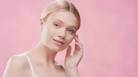 Young slim cute European woman with long light hair in ponytail in white crop top touches jawline turning her body and looking at the camera against pink ripple background | Skincare product ad