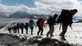 Young climbers in warm jacket, helmets and special glasses moving up on a mountain slope one by one in a row. Clip. People rising on snow covered slope on the background of many mountains and blue