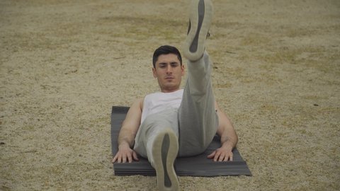 Muscular man doing bicycle style abdominal exercise, man in white tank top and gray tracksuit exercising, short hair man training in park, burning abdominal fat, man raising and lowering legs lying on