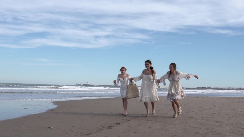 Three friends walking along the seashore in white dresses on a sunny day.