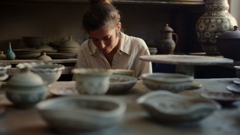 Closeup focused woman making ornament on clay pot in pottery. Smiling ceramist doing handcraft in workshop. Portrait of excited female artist drawing ornament in studio.