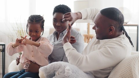 African family sitting on home sofa together, afro dad mature father man shows little daughter black child play game on fingers makes triangle with hands gesture baby sits with mom repeats after daddy
