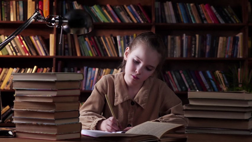Thoughtful schoolgirl writes home task in paper notebook sitting at table with book stacks and black lamp against large bookshelves at night
