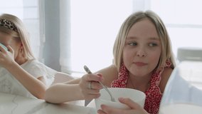 Two children eat a healthy breakfast in the kitchen in the morning. Happy Caucasian sisters enjoy granola or muesli and milk into a bowl. Slow motion 4K UHD video shooting 50 fps