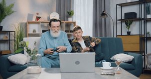 Good-looking happy carefree friendly bearded senior granddad and his grandson having fun together while playing video games on laptop and giving high five when win the game