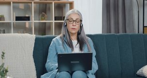 Attractive confident successful grey-haired senior woman in headset sitting in front of laptop during video conference and discussing report with charts with coworkers