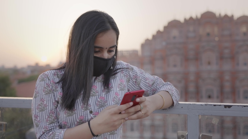Close shot of a young beautiful Indian female in traditional cloths wearing a protective face mask standing and using a mobile phone to type a text message against the Hawa Mahal Jaipur amid COVID 19 Royalty-Free Stock Footage #1070849179