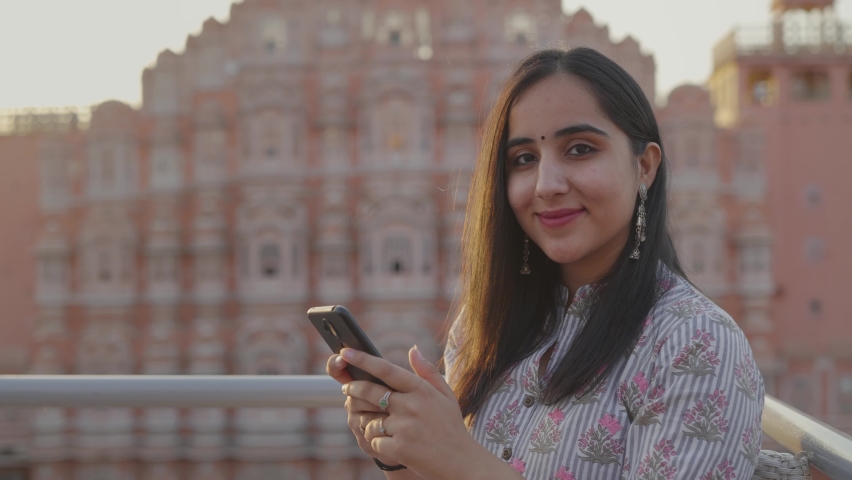Close view shot of a young beautiful Indian woman in traditional clothes against the historic Hawa Mahal, Jaipur in the background using a mobile phone and also smile looking at the camera  | Shutterstock HD Video #1070849200