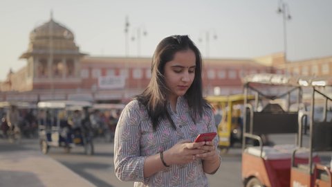 close view shot of young beautiful Indian female in the traditional dress using a mobile phone to type a text  message while walking on a footpath alongside busy road traffic and market place