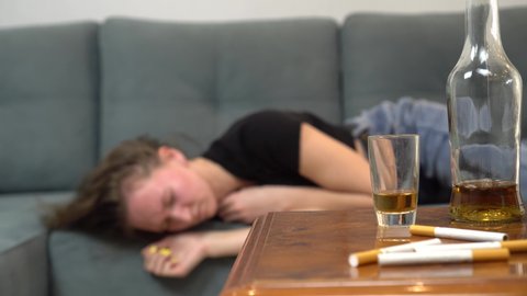 a drunk woman or girl is lying on the couch after a violent drinking session, next to her empty bottles, glasses and shot glasses. She's suffering from a hangover. Female alcoholism.