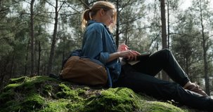 Young caucasian woman having video call from her laptop while resting in forest on a backpack. Concept of remote job, 5G internet connection and talking with friends. 