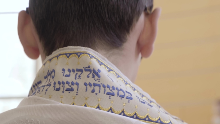 Jewish boy with Talit at a Bar Mitzvah. Kid with yarmulke Kipa having his ceremony at a synagogue in Israel. led by his family, Hebrew Torah words written on Talit, Tefillin. Judaism reform ceremony   Royalty-Free Stock Footage #1070862652