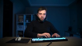 Bearded man working online at home on the computer at night in a room with blue light, looking at the screen with a serious face.