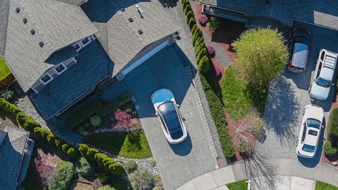 Aerial view of a white car driving through a suburban neighborhood arriving at home; drone follow shot
