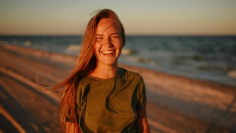 happy woman in khaki t-shirt, clothes and hair fluttering in the wind. young thin beautiful blonde woman laughs smiling rejoices having fun at sea on the beach at sunset in front of camera. close view