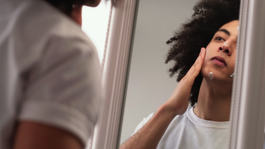 Man skincare. Facial moisturizing. Cosmetic product. Confident handsome guy with long curly hair applying smearing face cream looking in mirror. | Shutterstock HD Video #1070882134
