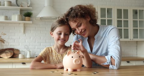 Woman her daughter sit at table in kitchen smile look at camera, little girl puts coins in piggy bank, mom teaches kid value of money, save finance, think about future, manage personal savings concept