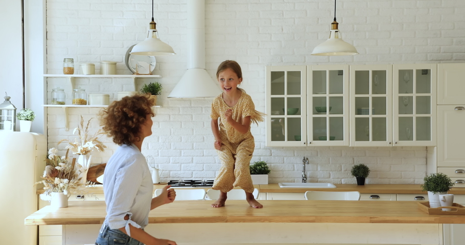 Vivacious little cute girl perform cool funky dances on dining table having fun together with young carefree mother in cozy modern kitchen on weekend. Home hobby, active quarantine with kids concept Royalty-Free Stock Footage #1070885560