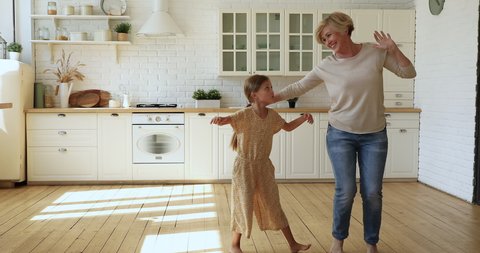 Cheerful active 60s granny little girl adorable grandkid listen music dance barefoot on warm wooden floor with underfloor heat system in cozy kitchen, full length. Funny activity, home hobby concept