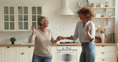 60s woman dancing with grown up daughter in kitchen, celebrate event holiday enjoy active weekend hobby, fun together. Family bond, tenant, healthy retiree and adult children, happy motherhood concept