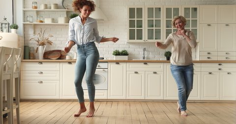Vivacious young woman dancing barefoot in kitchen with lively pensioner elderly mother, family having fun at modern home, enjoy life and active weekend together, listen music funny moving feel happy